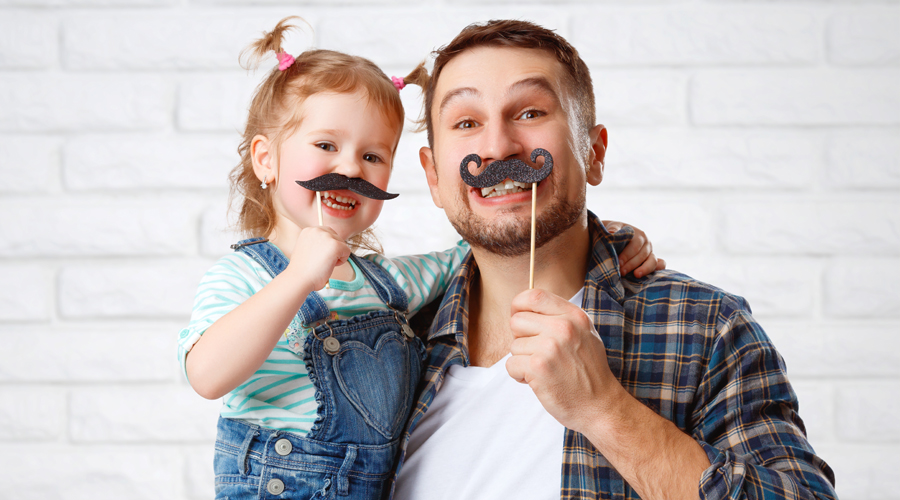 Smiling father and young daughter posing indoors against a white brick wall, each holding playful fake mustache props on sticks.