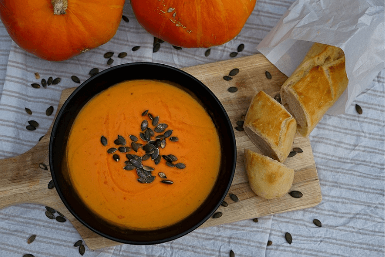 A bowl of pumpkin soup with pumpkin seeds on top and some pumpkins and bread can be seen in the side of the image