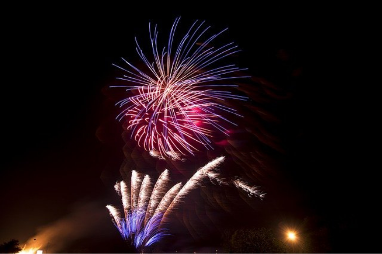 Pink and purple fireworks against the night sky and the tip of flames of a bonfire can be seen at the bottom of the image