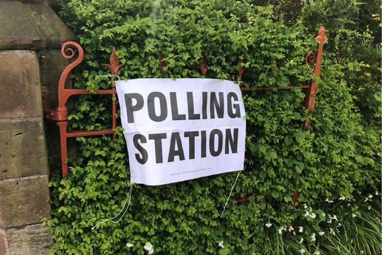 A white sign with black text reads 'polling station' - the sign is fixed to a gate with lots of greenery behind it.