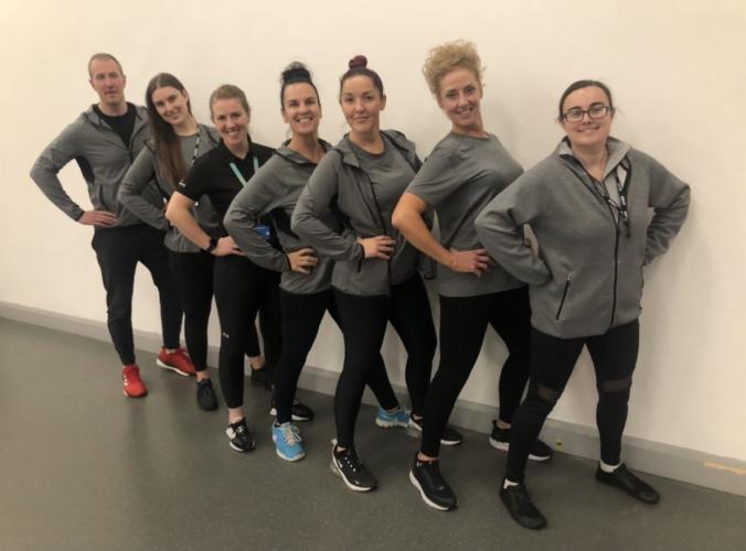 Staff from Wirral's Weight Management team with Lucy Holmes from One Wirral CIC (third from left). Seven people are standing in a line against a blank wall, with their hands on their hips and smiling.