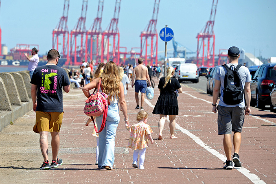 In the foreground, people's backs as they walk along New Brighton Prom in summer. In the background, large red cranes from Liverpool docks