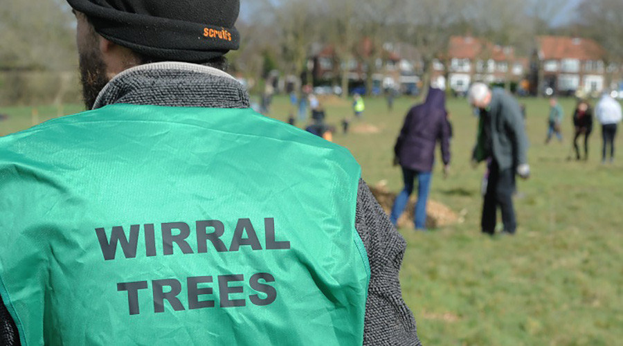 In the foreground, the back of a volunteer wearing a green 'Wirral Trees' tabbard, whilst in the background other volunteers plant trees