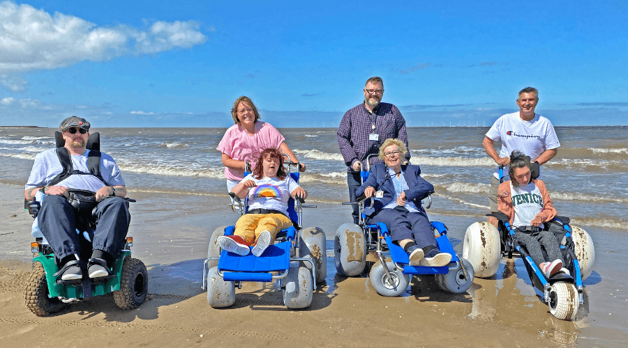 Group of people on New Brighton beach with the sea behind them, some in the new beach wheelchairs