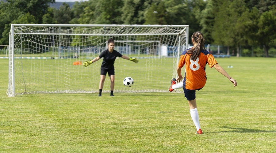 girl in orange football shirt kicking a ball towards a goal while goalkeeper in black football shirt attempts to save the shot