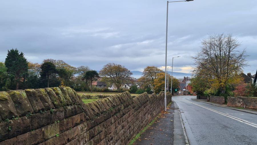 Front facing view when walking down to West Kirby. Stone wall on left hand side with fields, road on the right hand side and a view of the water and hills ahead.