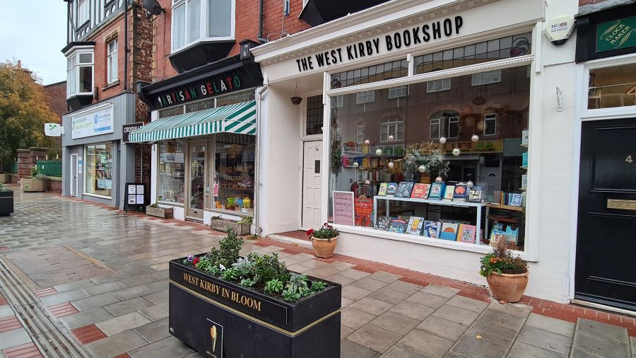 Image of a small section of row of shops including a bookshop and a gelato shop. A planter is in the front of the photo for In Bloom.