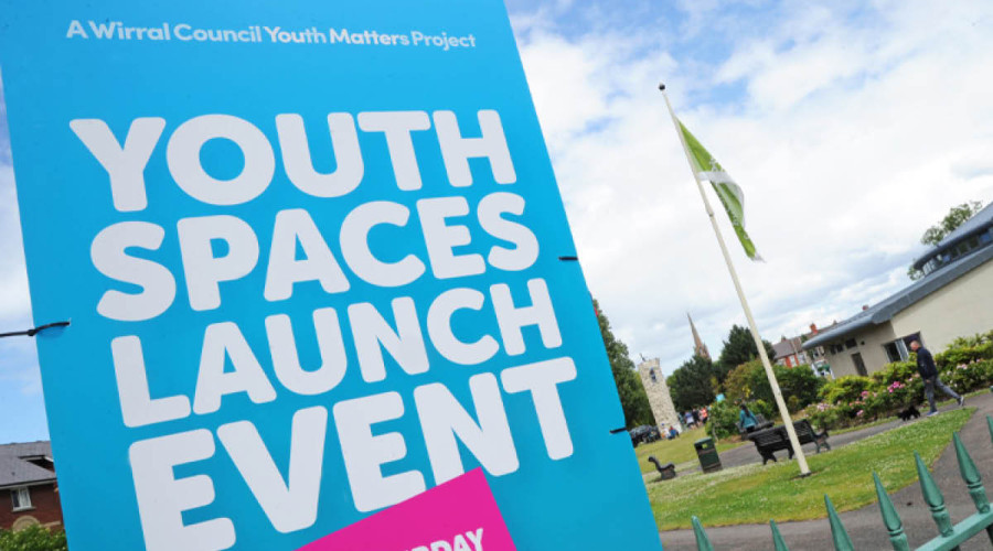 Background picture of a sunny sky over a Wirral park, in the foreground is a blue sign with white lettering reading 'Youth Spaces launch event'