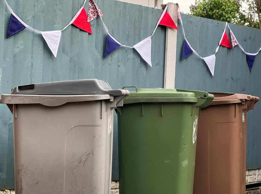 Grey, green and brown wheelie bins in a row in front of a blue fence with red, white and blue bunting attached to it.