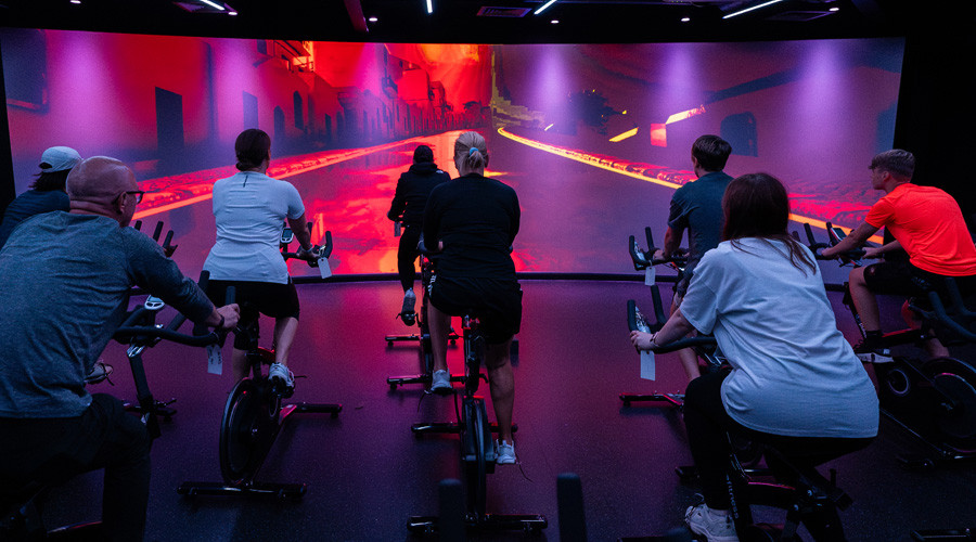 Group of people on exercise bikes in front of a floor to ceiling screen with pink visuals