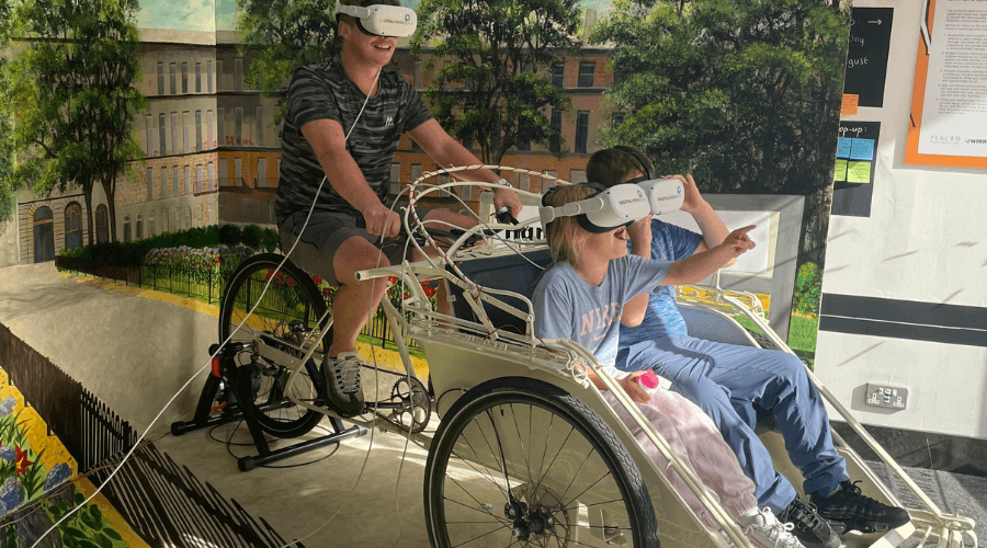 People enjoying the Virtual Reality experience. All are wearing Virtual Reality headsets. One person is cycling on a static bike and the two younger people in the front are sitting in a bucket seat and one is pointing at something in front of them that they can see through the headset.