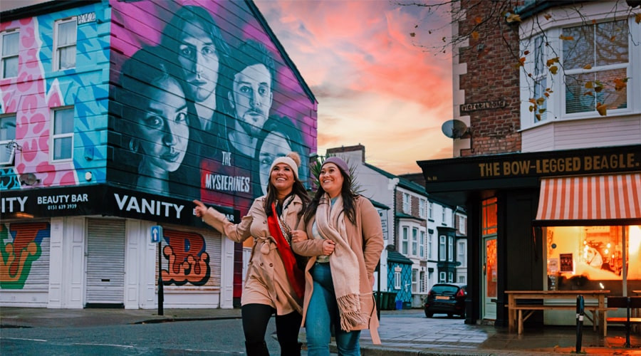 image of two friends crossing Victoria Road in New Brighton. In the background a mural of the band The Mysterines can be seen painted on the building