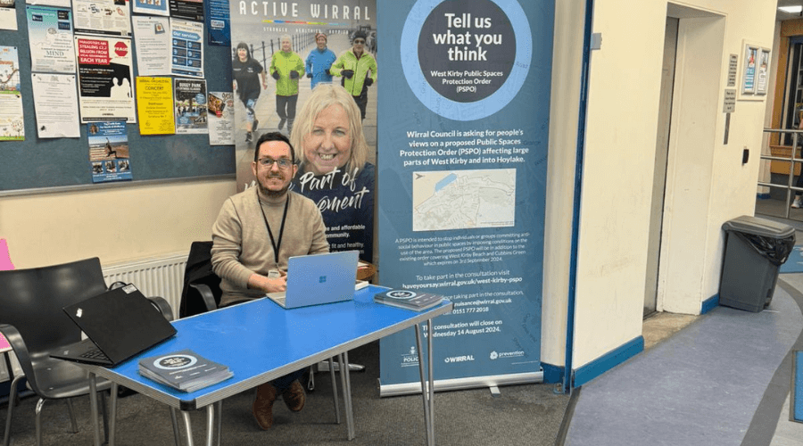 Person saitting behind a desk in the foyer of West Kirby Concourse, with a roll up banner behind him that says 'tell us what you think - West Kirby Public Spaces Protection Order (PSPO)' 