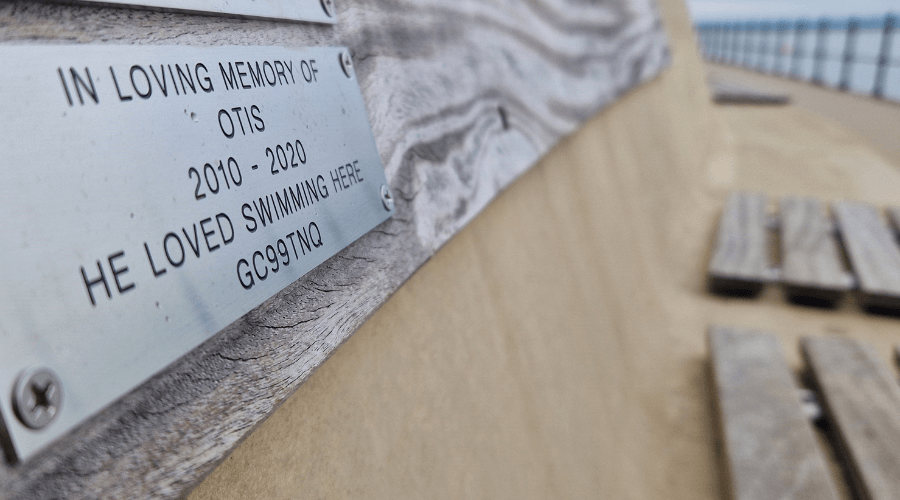 Close up of a memorial plaque on the West Kirby Flood Wall that says 'In loving memory of Otis 2010-2020 He loved swimming here' The plaque also features a code as the plaque is a geocache site.