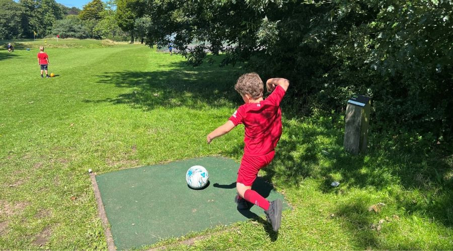 Child wearing a football kit kicking a ball on a golf course