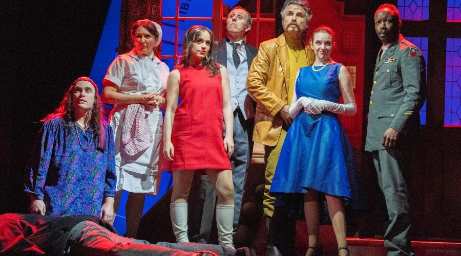 7 members of the cast of Cluedo stood over a body lying on the floor
