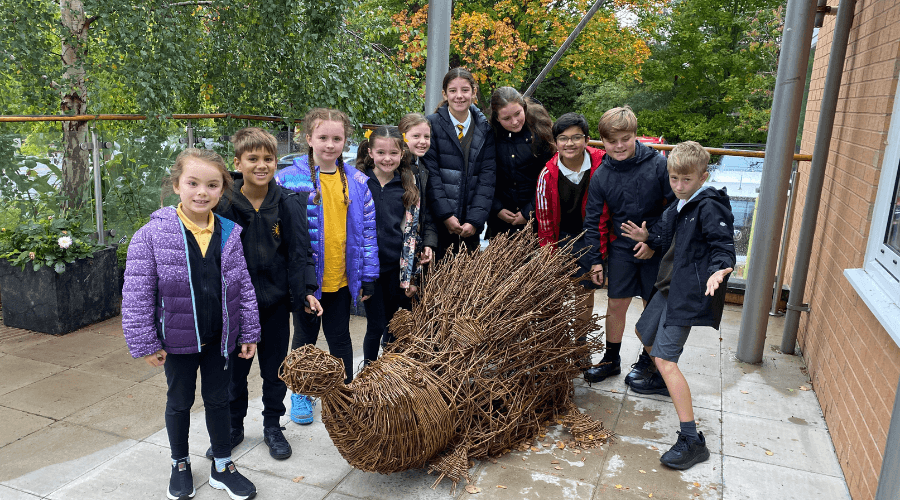 A group of school children gathered around a large hedgehog sculpture made out of willow