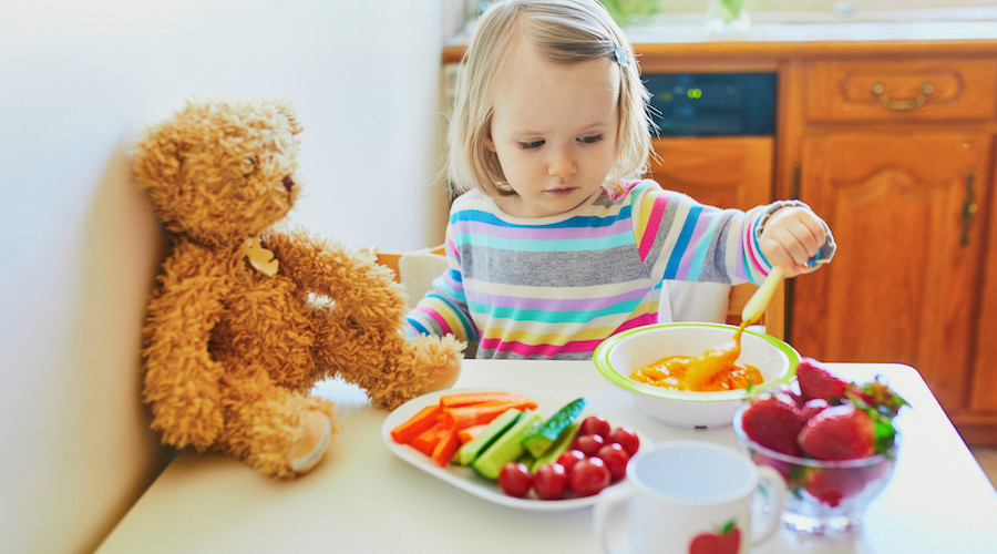 Toddler girl with blonde hair using spoon to get food from bowl, her teddy bear is sitting on table waiting to be feed and a dish of mixed fruit and vegetables are in front of her.