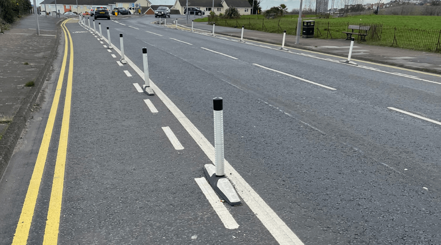 A section of road with a cycle lane that not only has a white line separating the road and the cycle lane but it supported by orcas that provide more of a barrier between them and yellow lines.