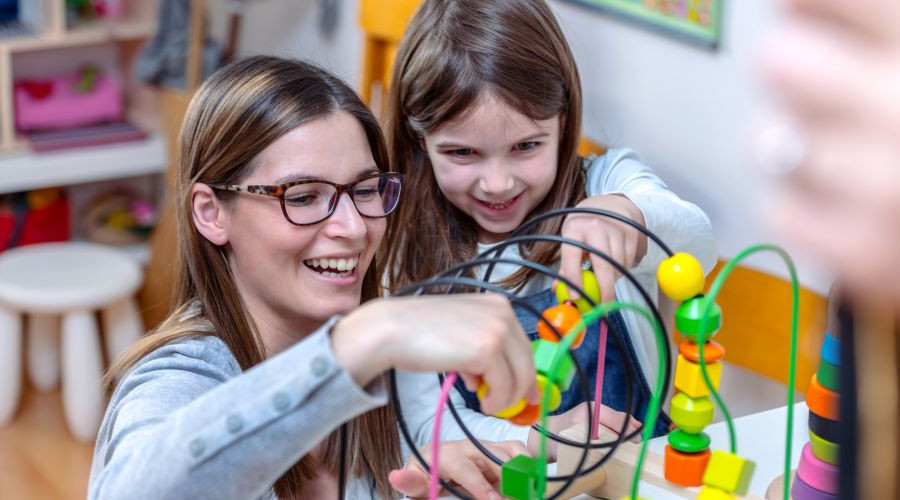 A woman with long brown hair in a grey jumper playing with a young girl with dark brown hair. They are both smiling.
