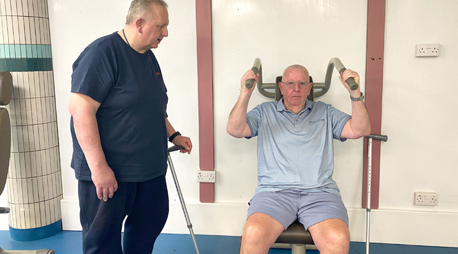 Barry is standing with his walking stick next to Rob who is sitting down on exercise equipment. Barry is showing Rob how to use the equipment.