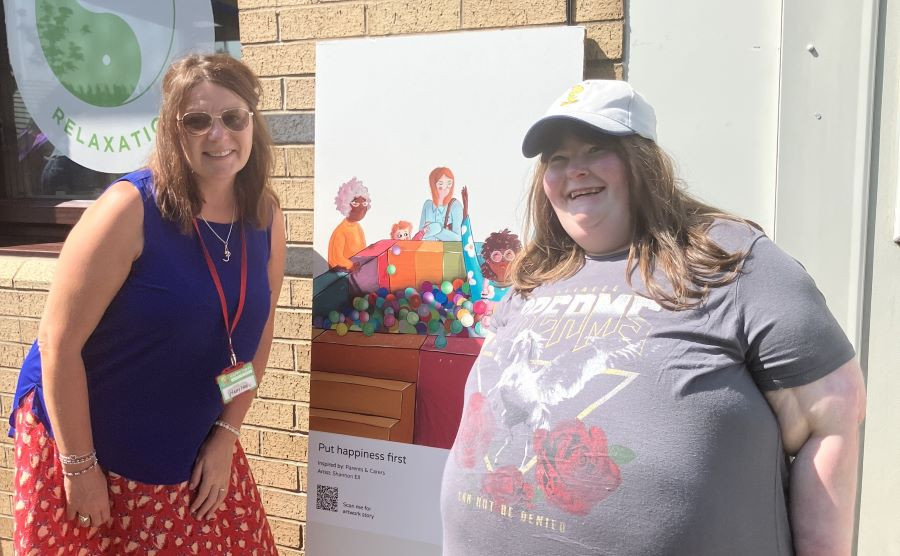 Two women standing next to an art board. One is wearing a grey t shirt and white cap and the other is wearing a blue vest top and red skirt. They are both smiling.