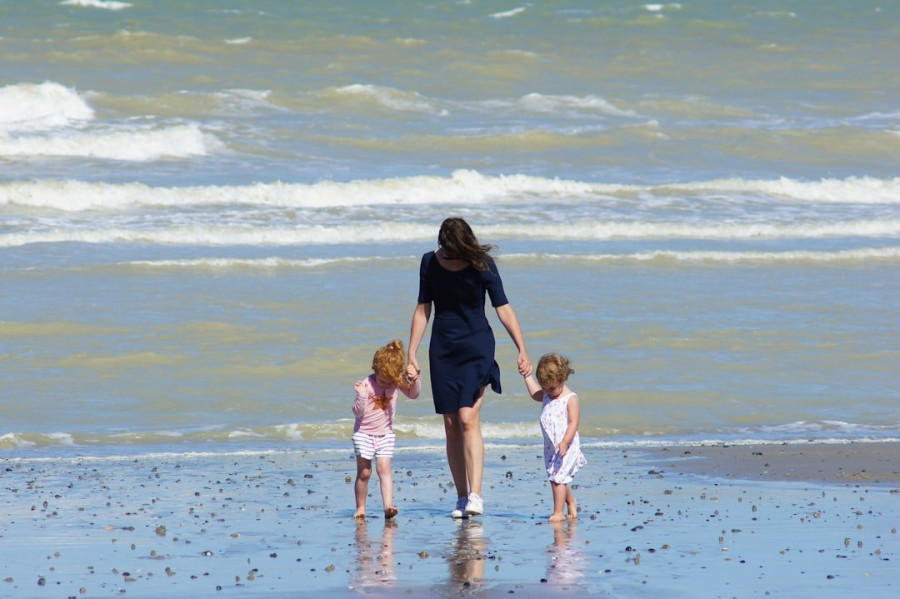 A woman in a blue dress is walking along a beach holding the hands of two children