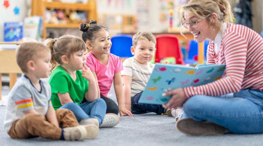 four nursery age children smile as a teacher reads to them from a book, they are sitting on the floor