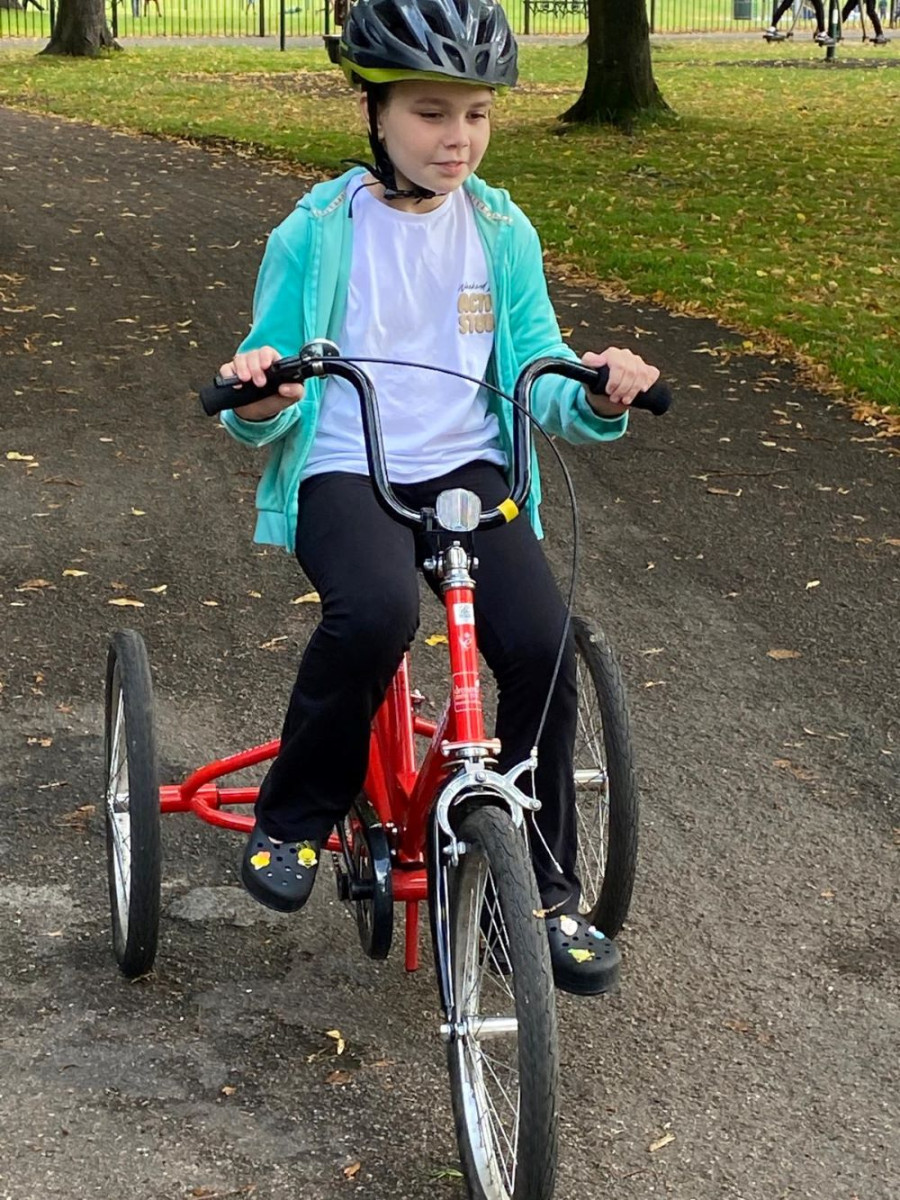 A young girl in a white t shirt and blue jacket  riding an accessible red tricycle on a park path, promoting inclusive cycling.