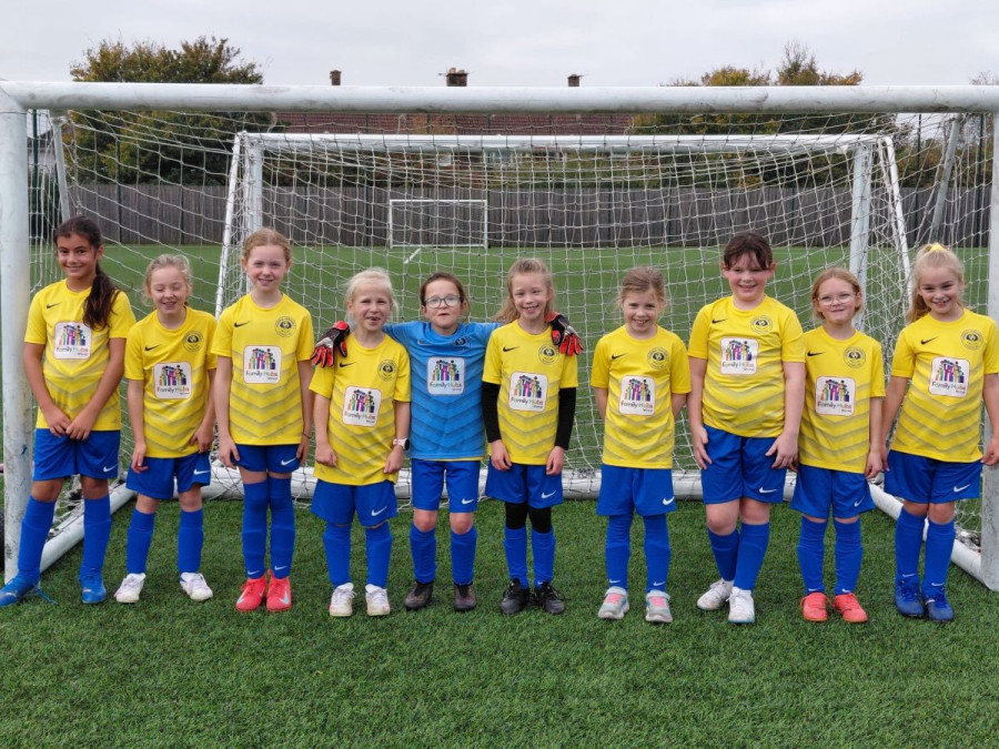 Ten young children in yellow and blue football kits standing in a line on a green pitch in front of a goalpost. One child in the middle wears a different kit, likely the goalkeeper. They are smiling and look ready to play. The image shows a local youth football team, highlighting community sports for children.