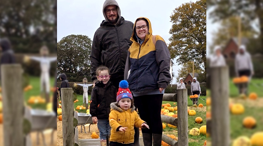 Four people standing on a wooden footbridge at a pumpkin patch. The group is dressed in warm, waterproof jackets and boots, with one child wearing a bright yellow coat and a colorful bobble hat. Pumpkins of various sizes are scattered across the grassy field and straw-covered paths in the background. Other visitors and wheelbarrows are visible among the pumpkins, with large trees and an overcast sky completing the autumn setting.