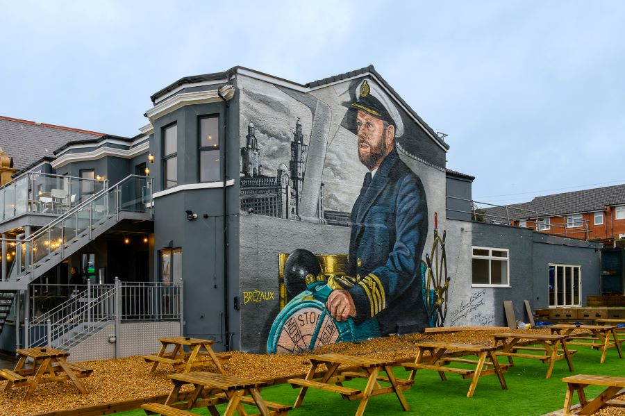A large mural painted on the side of a grey building, depicting ship captain Mike Lowe in a navy uniform holding a ship’s wheel. Behind the captain, the artwork includes a detailed black-and-white cityscape featuring tall historic buildings. The building has an outdoor seating area with wooden picnic tables on green artificial grass, and a staircase leading to an upper deck with string lights.