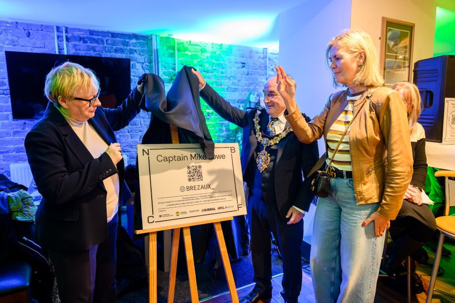 Angela Eagle, Cllr Bryan Kenny and Cllr Paula Basnett standing indoors during an unveiling ceremony, lifting a black cloth from a sign on an easel. The sign reads “Captain Mike Lowe” and includes a QR code, logos, and directional markers. The background features a brick wall and colorful lighting.