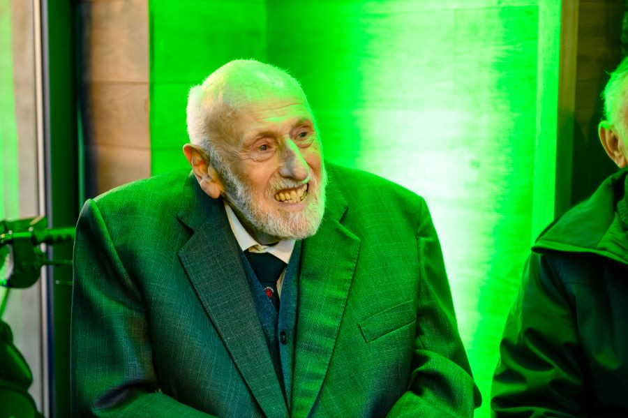 Captain Mike Lowe pictured wearing a dark green suit jacket over a layered outfit, seated indoors at the event against a bright green illuminated background. Part of another person in a dark coat is visible to the right.