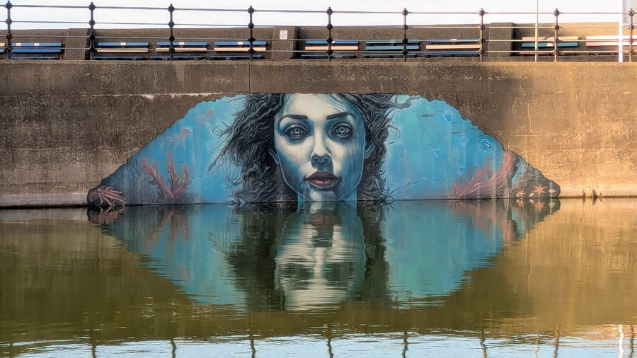 A mural painted on the side of a concrete bridge, depicting an underwater scene with blue tones, seaweed, coral, and marine life. The artwork is partially reflected in the calm water below, creating a mirrored effect. The bridge has a metal railing along the top and appears weathered, contrasting with the vibrant colors of the mural.