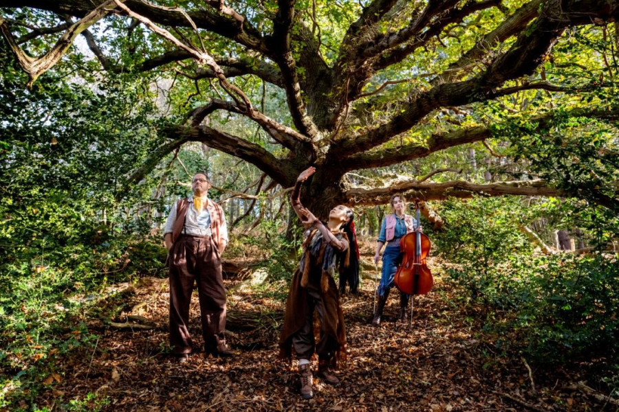A performer in woodland costume stretches an arm upward beneath the wide branches of an ancient oak tree, while two other performers stand nearby, one holding a cello. The group is surrounded by dense green foliage and dappled sunlight in a forest setting.