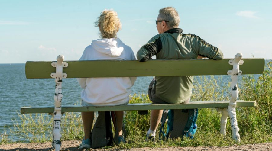Man and woman witting on a bench by a beach with their back to us