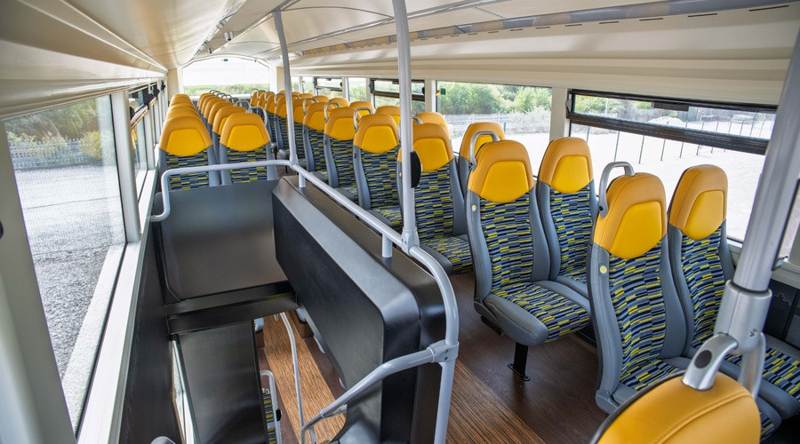 Interior of a double-decker bus showing rows of yellow and patterned seats, wide windows, accessible seating areas and a central staircase, viewed from the upper deck.