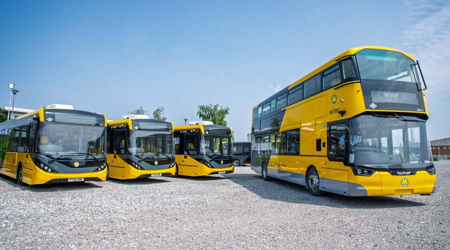 Fleet of Wirral's new yellow buses parked outdoors, including a double-decker and several single-decker vehicles, pictured in a depot under a clear blue sky.