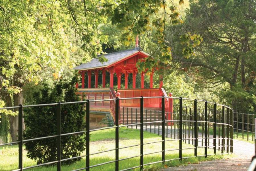 Birkenhead Park. The red swiss bridge is in the centre of the shot with the railings of the path in the foreground. It is a sunny day.
