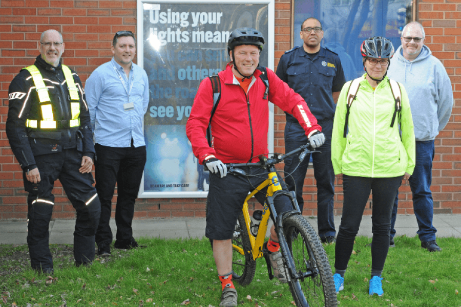 Group of people standing in front of a poster that has a picture of a bike and the words 'using your lights means that you can see and others on the road can see you too'
