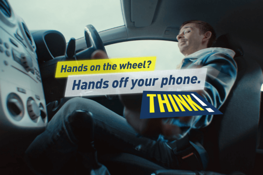 A man sitting in car and glancing down at his phone to see text messages on his phone - there is some text across the image that reads 'Hands on the wheel? Hands off your phone.'