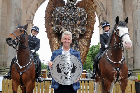 In the foreground, a person holding a silver anti-violence plaque, flanked by two police mounted on horseback. In the background, the Knife Angel statue