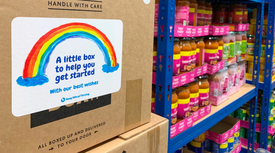 Wirral Summer Food support for children box with rainbow sticker in foreground and non-perishable food stacked on shelving in background