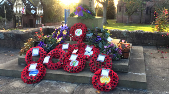 Multiple wreaths laid at the foot of stone cross in Thornton Hough with sun shining through trees