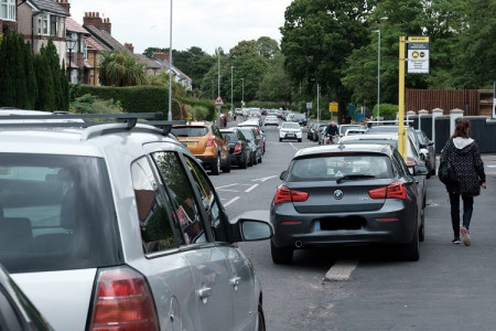 image of Bebington street with cars parked partly on pavements and in bus stop as a person walks past