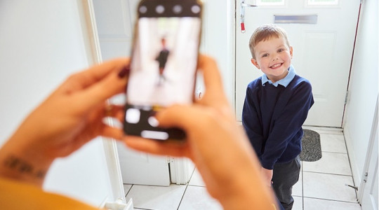 A smiling child in school uniform by the front door as a parent takes a photo on their phone