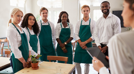 Stock photo showing a team of hospitality staff in white shirts and green aprons receiving instructions from a supervisor