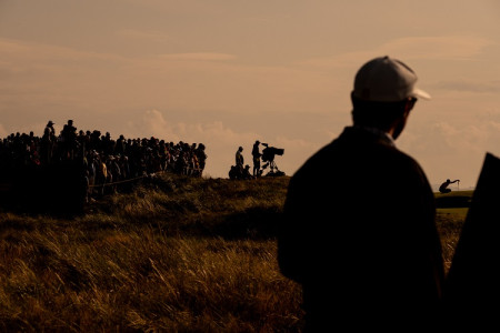 Silhouette of a golf fan in the foreground and a tv crew further away