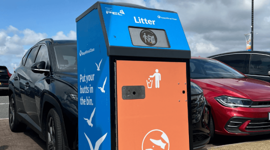Close up of an enclosed bin in orange and blue colours. You can see a row of cars in the background as the bin is on the pavement of the promenade in New Brighton and the bin has a solar panel on the top with a message on the side saying 'Put your butts in the bin'